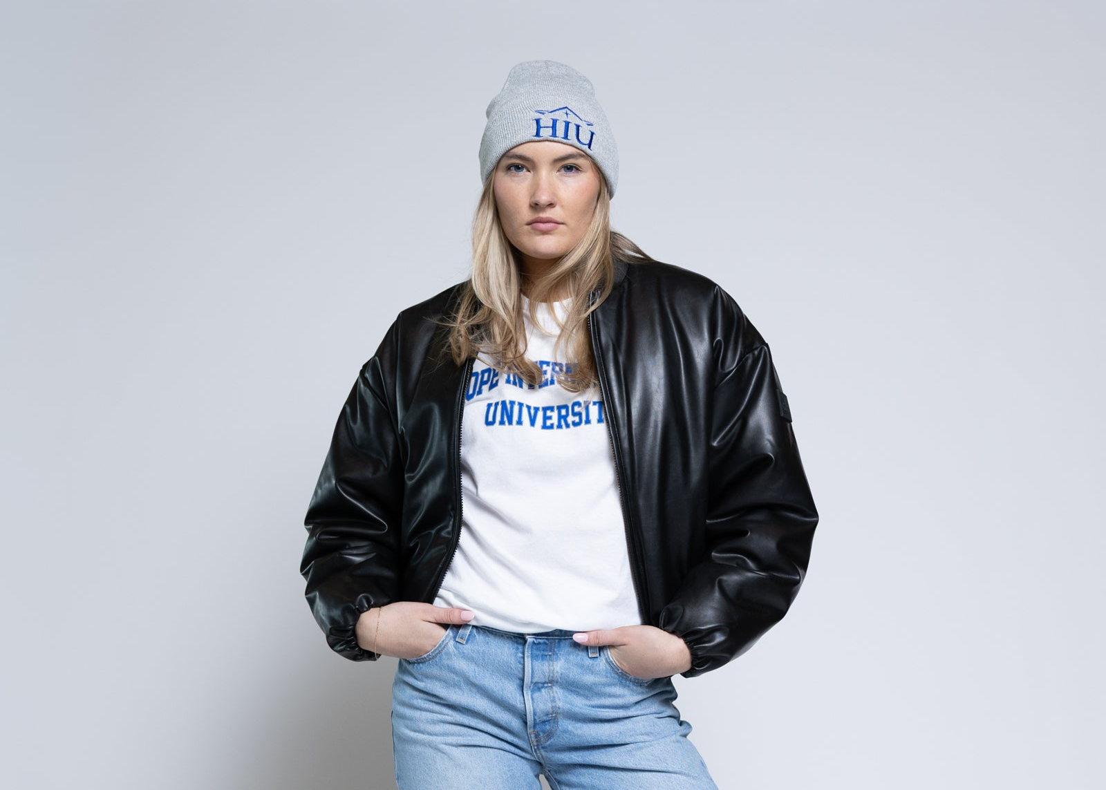 Woman wearing a gray hat, black leather jacket, white t-shirt with text, and blue jeans on a plain background